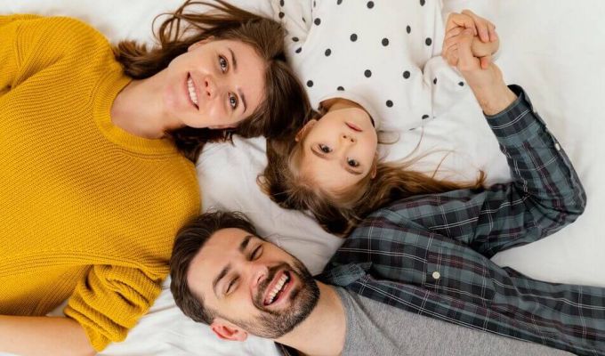 Zdjęcie typu "flat lay" przedstawiające uśmiechniętą trzyosobową rodzinę leżącą na białej pościeli. Po lewej stronie kobieta w miodowym swetrze, na dole mężczyzna z brodą w kraciastej koszuli, a pomiędzy nimi mała dziewczynka w białej bluzce w kropki, trzymająca tatę za rękę. Wszyscy patrzą w stronę obiektywu.