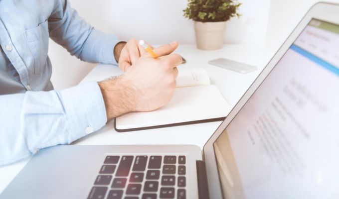 Zdjęcie typu zbliżenie przedstawiające osobę w błękitnej koszuli, która pisze żółtym ołówkiem w notesie. Obok na białym biurku stoi otwarty laptop, a w tle widać małą roślinę w doniczce.